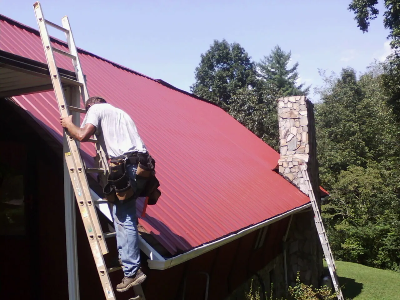 Roofer on a ladder beside a red metal roof and stone chimney — Soffit & Fascia Repair by Roofing Brothers in Allgood